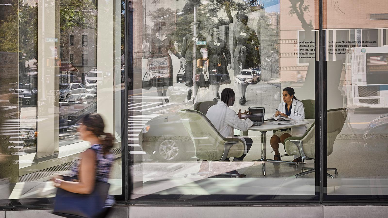 Exterior street view of School of Nursing, showing a passing pedestrian and employees meeting inside