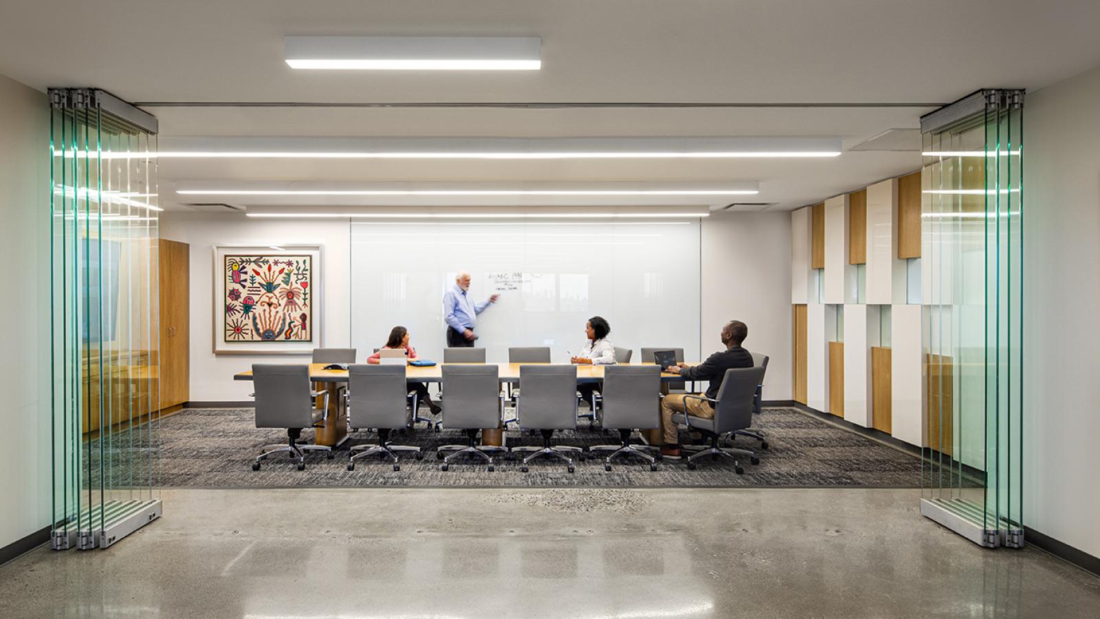 A man standing at a white board in a conference room with three people sitting around a table