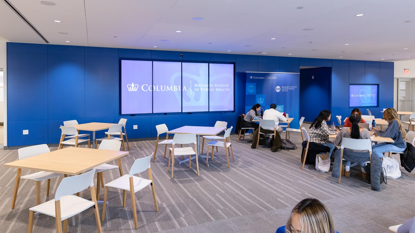 a lounge with a blue wall and carpeting features square wooden tables with people sitting around them and a large digital screen on the wall.
