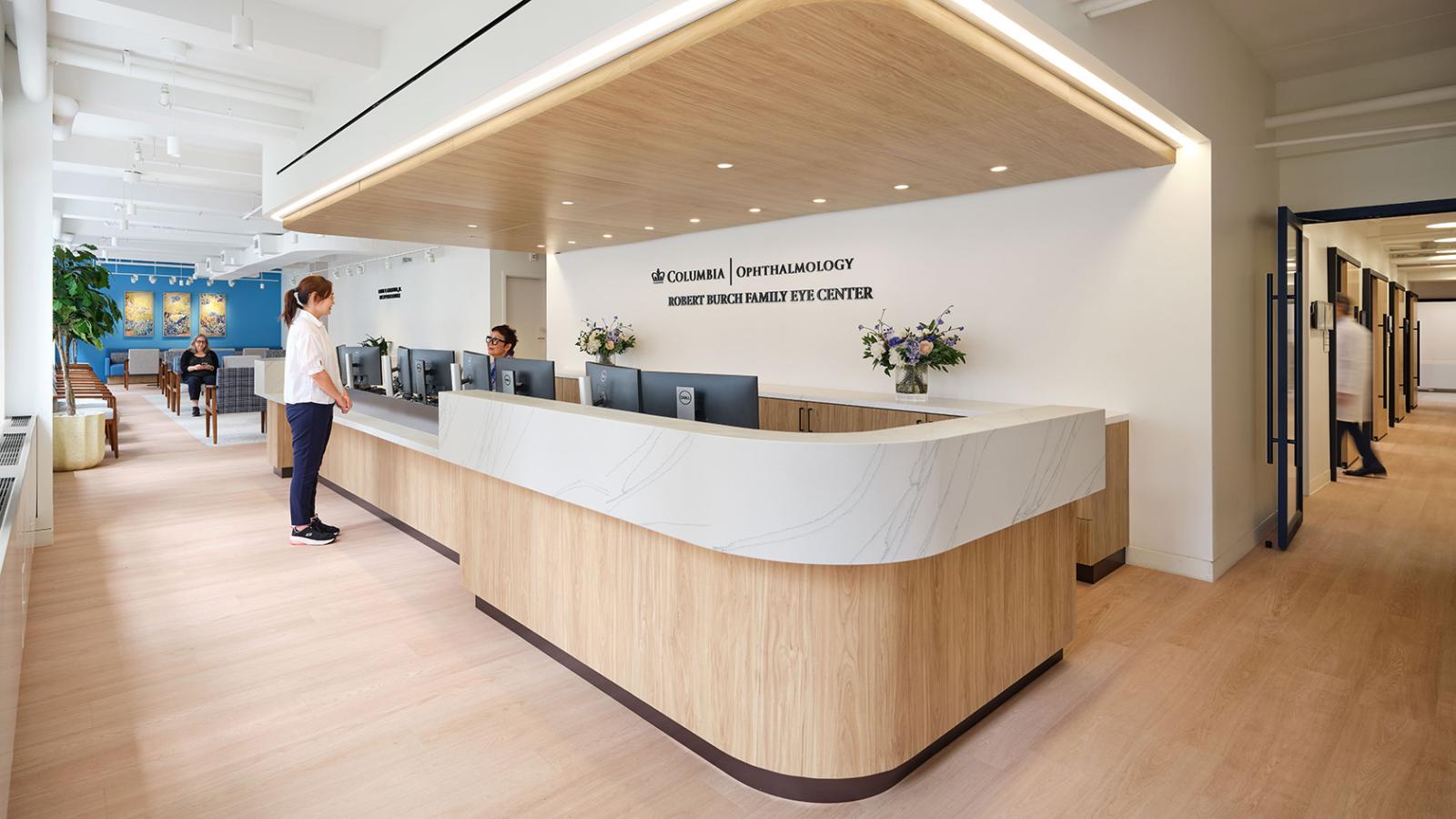Reception desk at the Robert Burch Family Eye Center with wood and marble finishes, staff assisting a patient, and a seating area in the background.