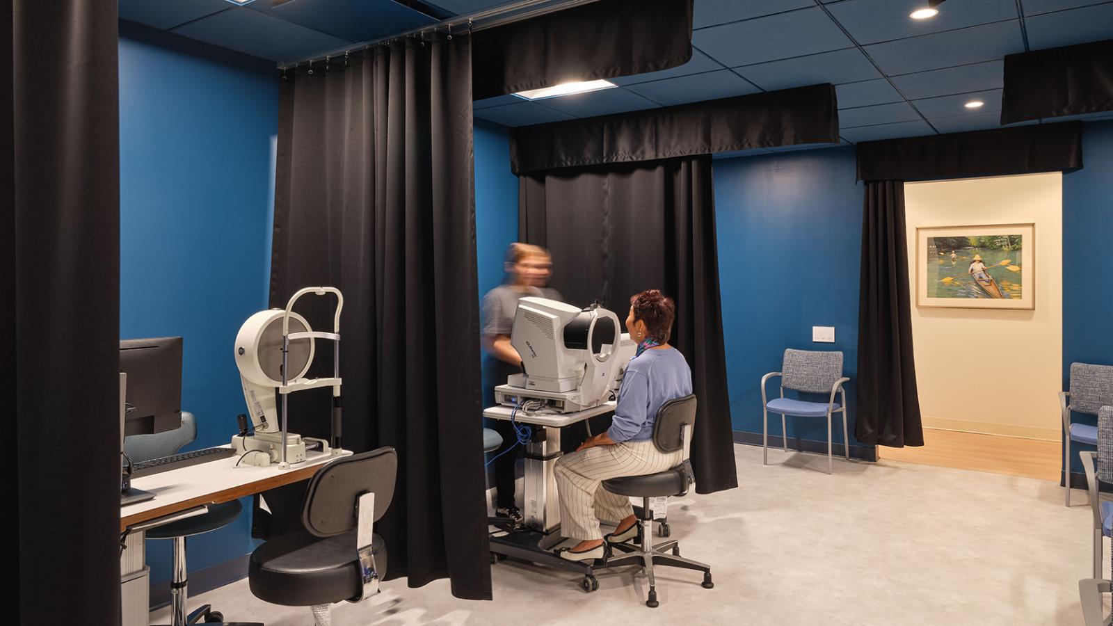 Patient seated at diagnostic imaging equipment in a darkened blue room with privacy curtains and clinical furnishings.