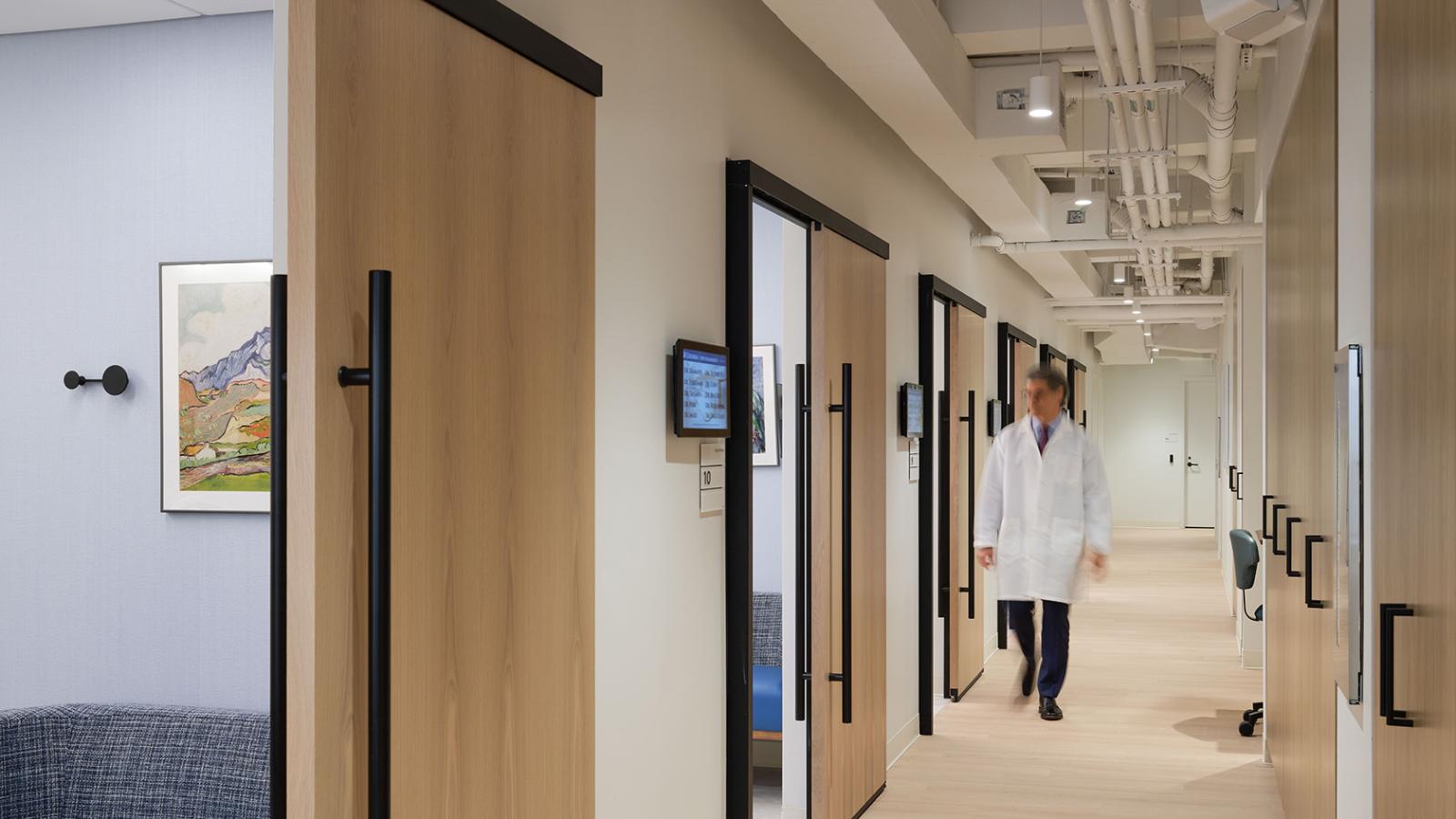 Corridor with sliding wood doors to exam rooms, overhead utilities, and clean modern finishes, with physician walking down the hall.