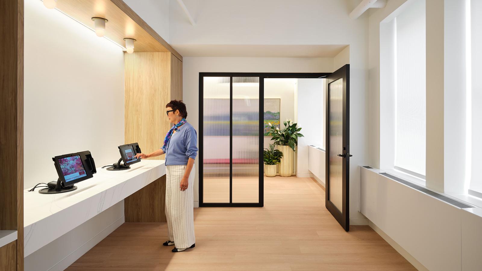 Patient using electronic check-in kiosks in a bright alcove with wood accents, modern lighting, and adjacent waiting area.