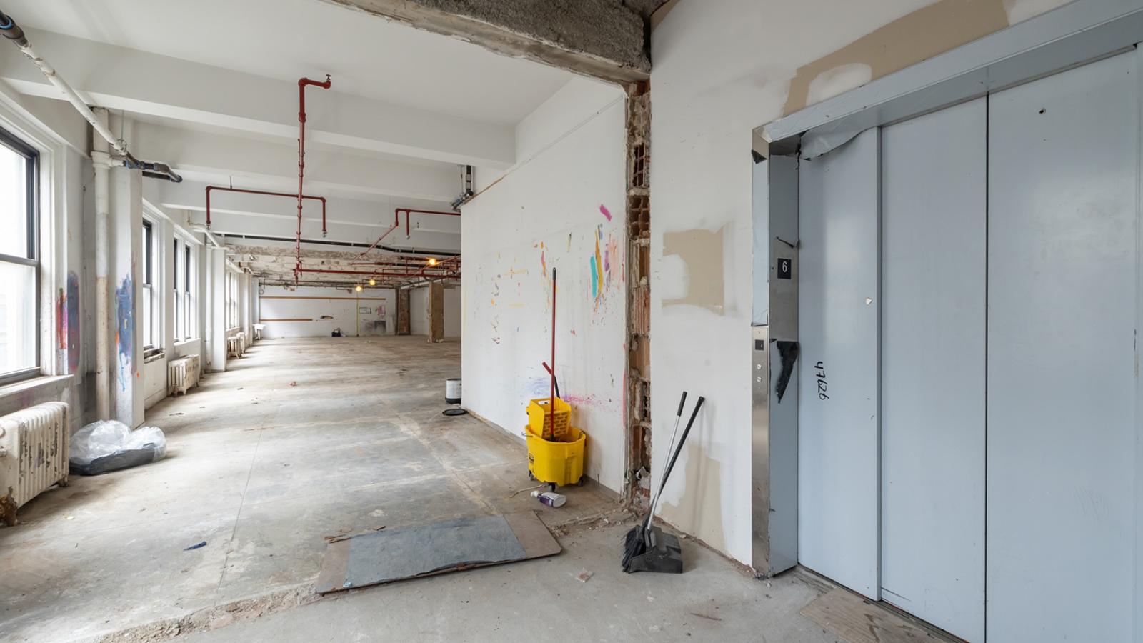 Partially demolished interior near an elevator lobby with exposed walls, pipes, and construction materials.