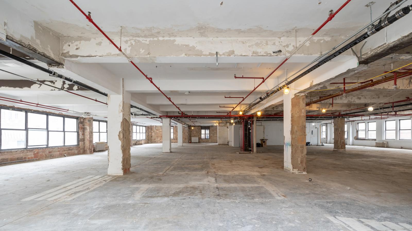 Empty floor under renovation with exposed concrete columns, ceiling beams, and visible piping.