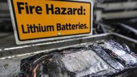 A yellow sign with black letters that spell Fire Hazard: Lithium Batteries. In front of it lies a burnt cell phone